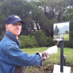 Masculine person in nature. They are wearing a baseball cap and jean jacket over a flannel shirt. They are holding a paintbrush up to a painting of the background landscape. They are also looking at the camera and smiling.
