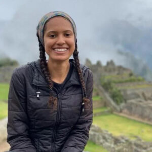 A woman wearing her brown hair in braids, a headband, and a black puffy jacket smiles in front of historic ruins.