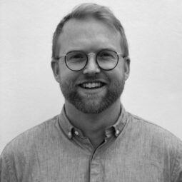 A man with short hair, a short beard, and glasses stands in front of a blank background, smiling at the camera.