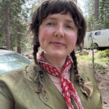 A person with brown braids and a red handkerchief stands in front of a forest smiling.