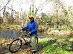 A man in a blue jacket and bike helmet standing with a bike in front of a landscape with a pond and trees.