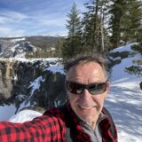 A person wearing a red plaid shirt and sunglasses taking a selfie with snowy mountains in the background.
