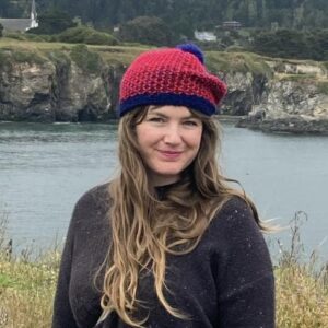A woman in a black sweater and red hat smiles in front of a body of water and cliffs.