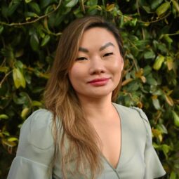 Yensing stands smiling in front of a background of green leaves, wearing a green shirt with long light brown hair over one shoulder.