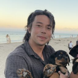 Matt on the beach smiling in front of a sunset, holding a puppy.