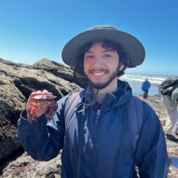 Alejandro smiling on a beach, wearing a sun hat and holding a crab