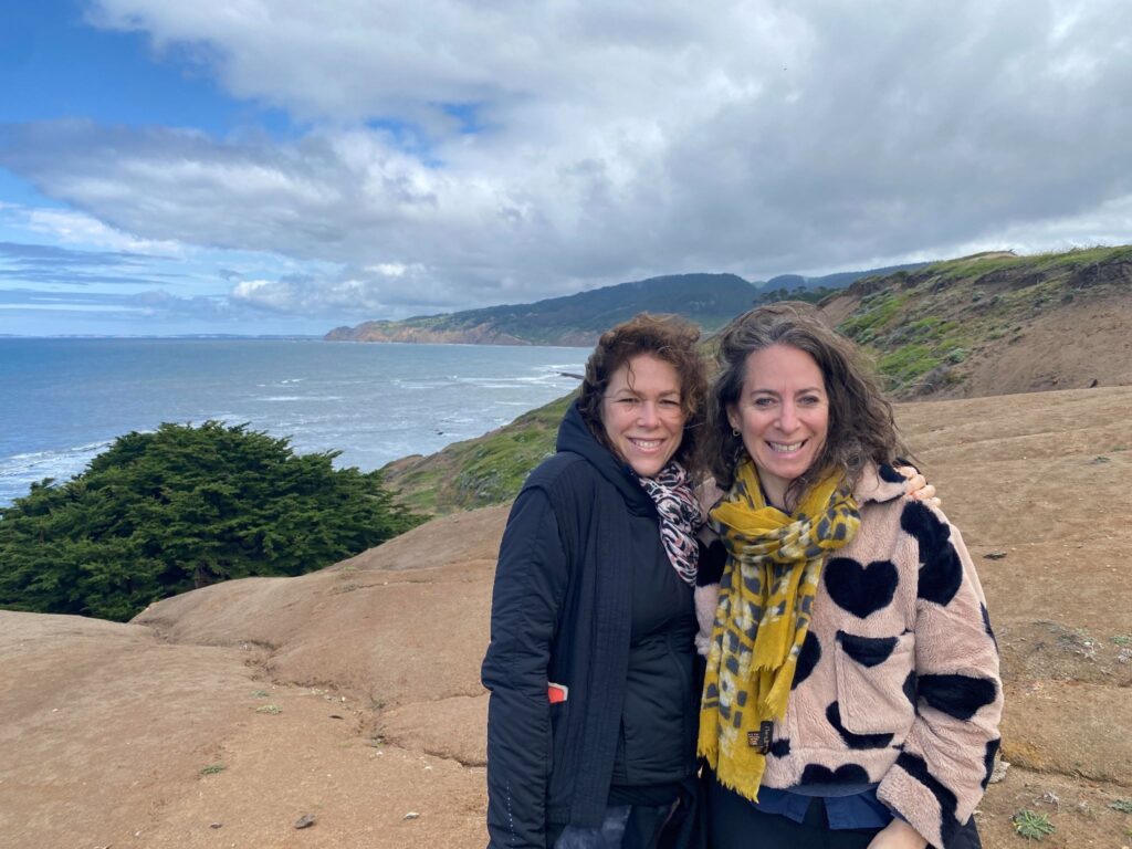 Two women smile at the camera with a coastal view in the background.